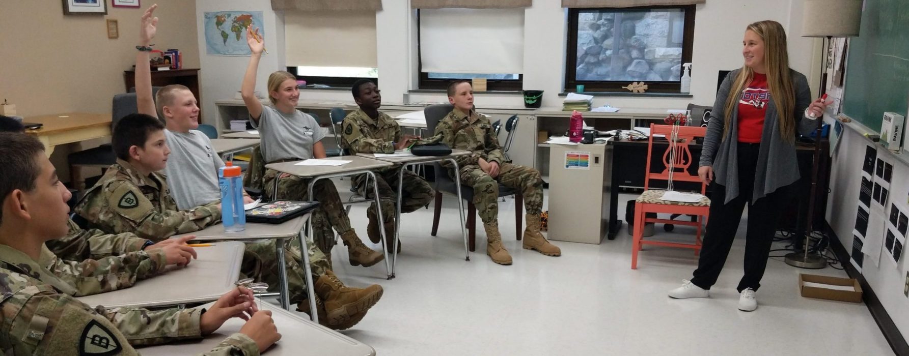 Cadets in Army camouflage uniforms raising hands in classroom with female teacher and world map display