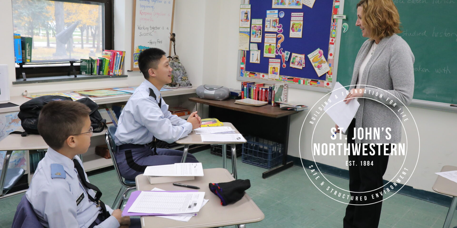 Cadets in blue dress uniforms receiving instruction from teacher in small classroom setting