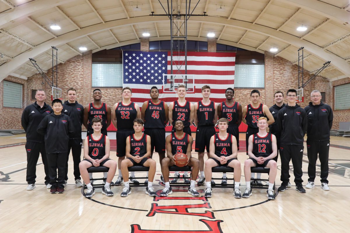 Varsity basketball team in black and red SJNA jerseys posed in gymnasium with American flag backdrop