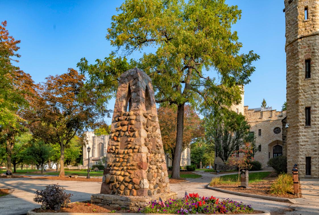 Stone cairn monument with statue centerpiece near historic stone tower building and chapel on tree-lined campus