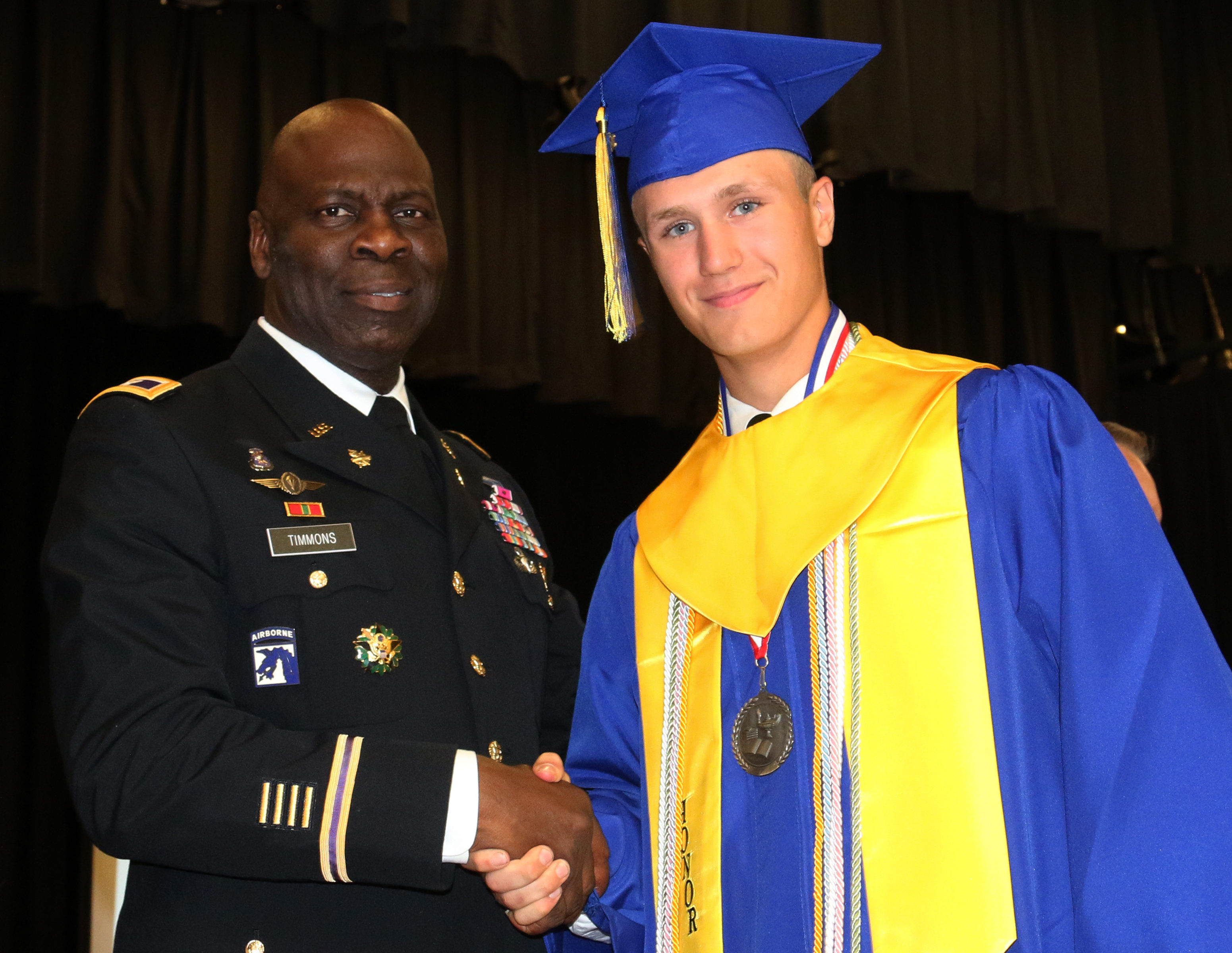 Army officer in dress uniform shaking hands with graduate in blue cap and gold gown