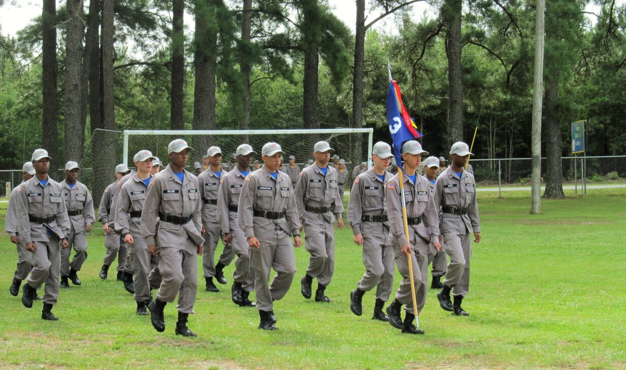 Cadet platoon in gray uniforms marching with guidon flag on grass field near pine trees