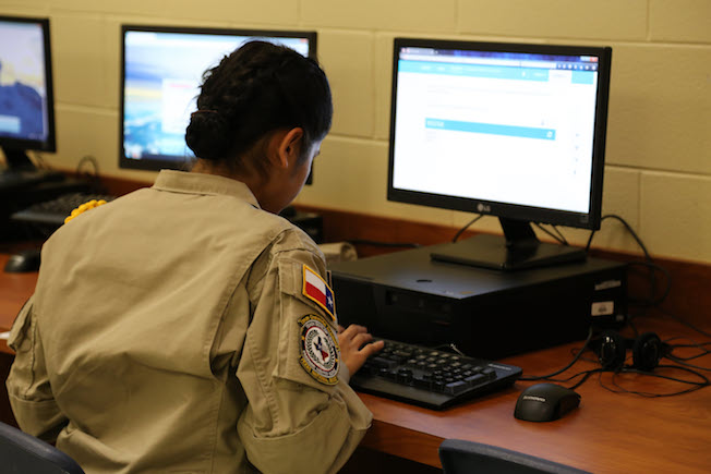 Female cadet in tan uniform with Texas patch works at desktop computer in classroom lab