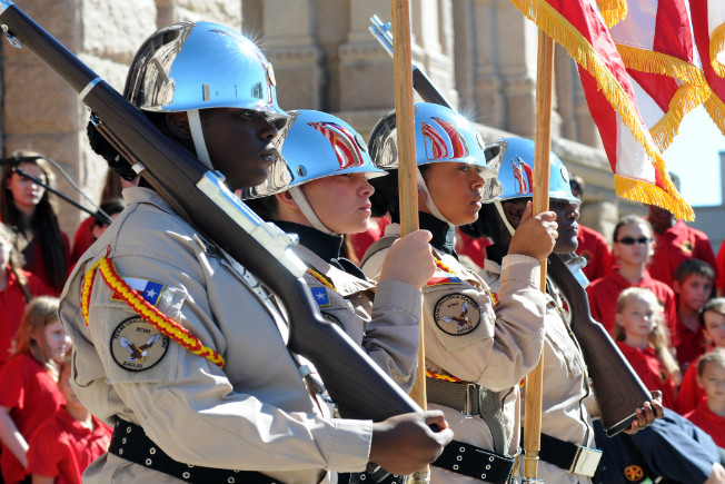 Color guard cadets in blue helmets carry rifles and flags during parade with spectators in background
