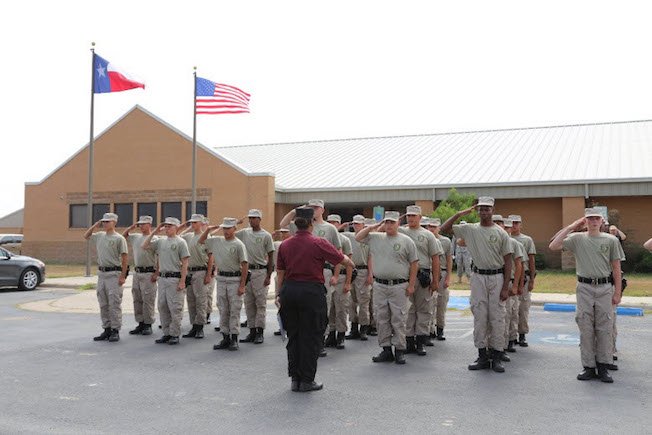 Cadets in tan uniforms salute in formation on campus with Texas and American flags flying overhead