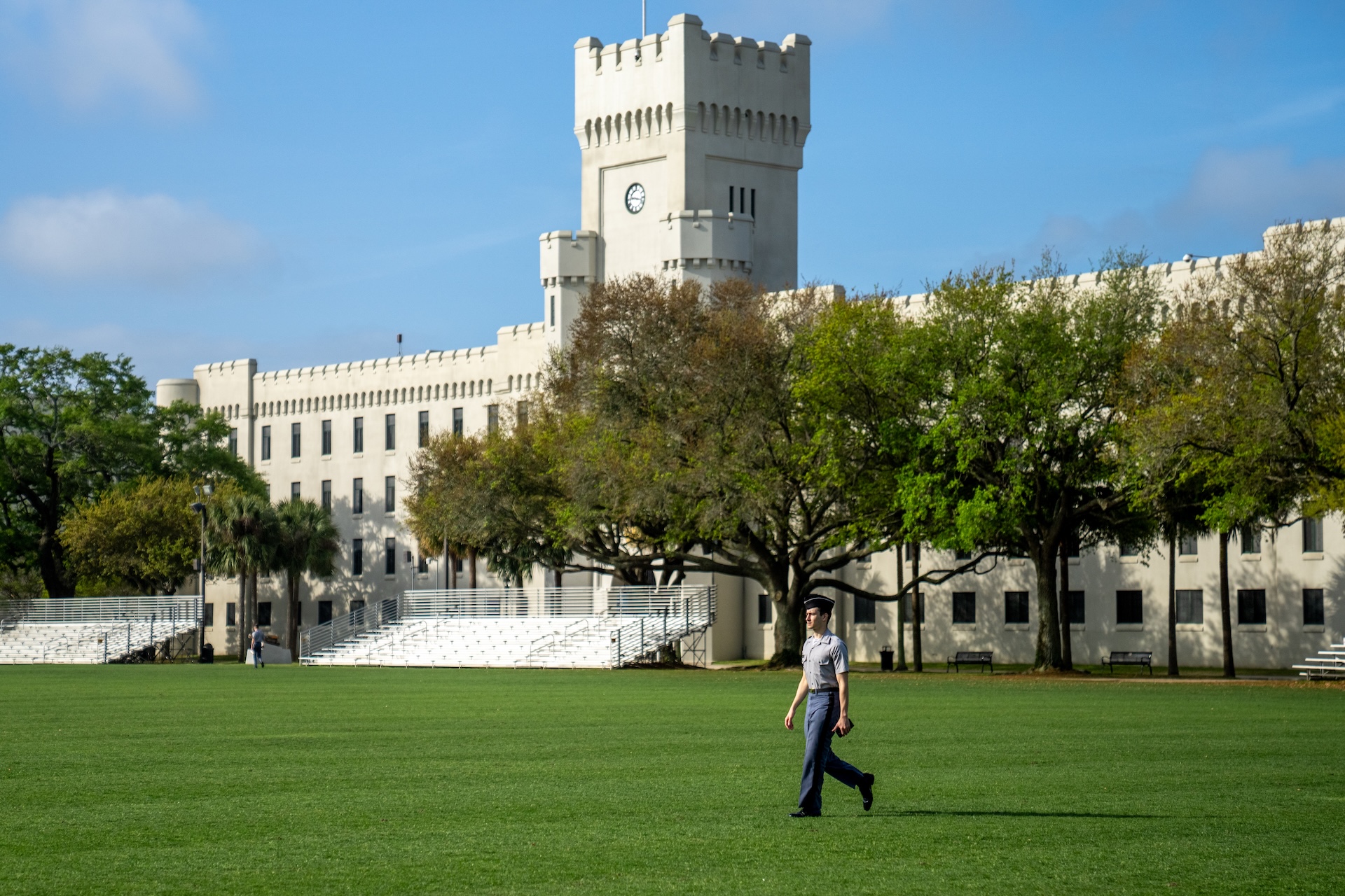 Cadet in gray uniform walking across the green parade ground with Padgett-Thomas Barracks and clock tower behind