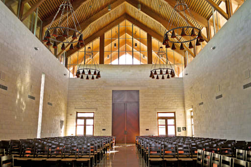 All Saints Chapel interior with vaulted wooden ceiling, iron chandeliers, limestone walls, and wooden pews