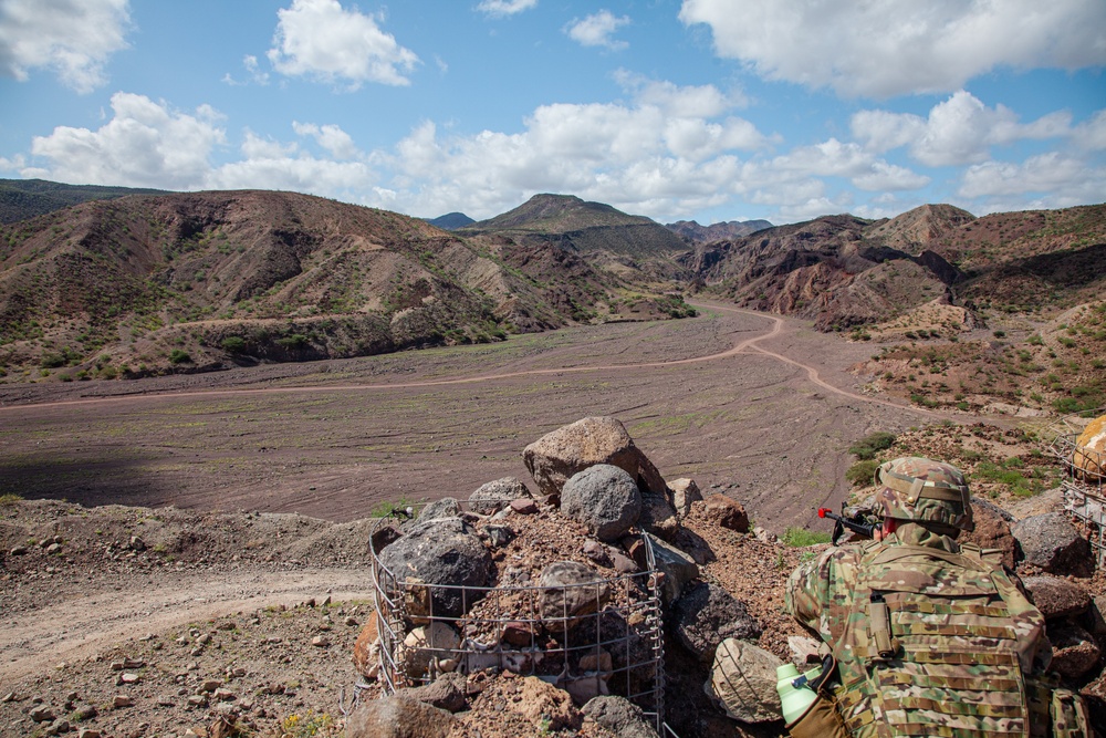 Airman in camouflage at observation post overlooking desert mountain valley terrain