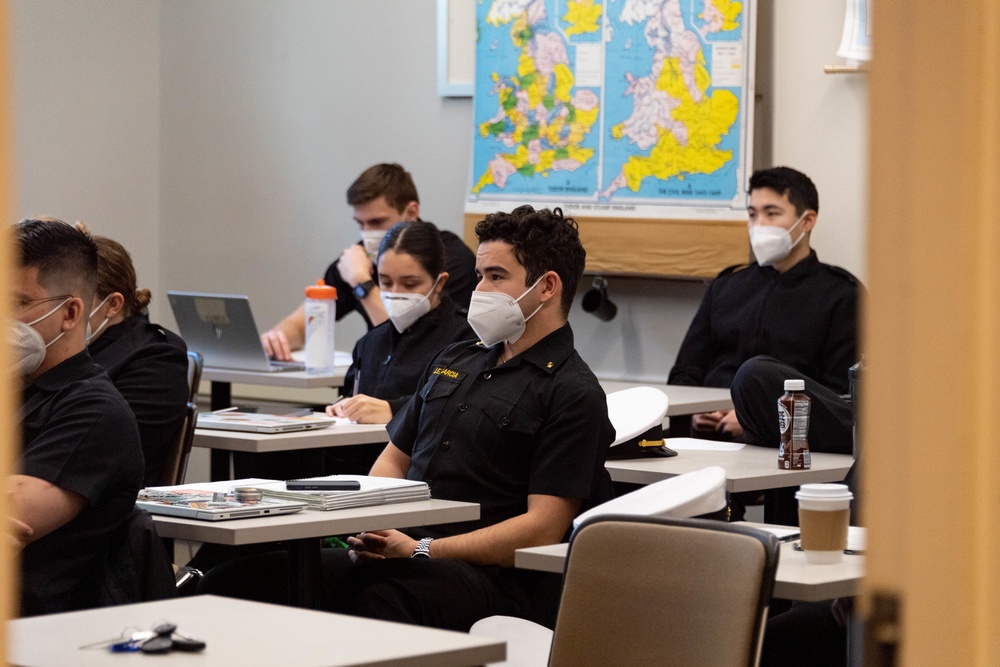 Midshipmen in dark blue uniforms studying in classroom with world maps on wall