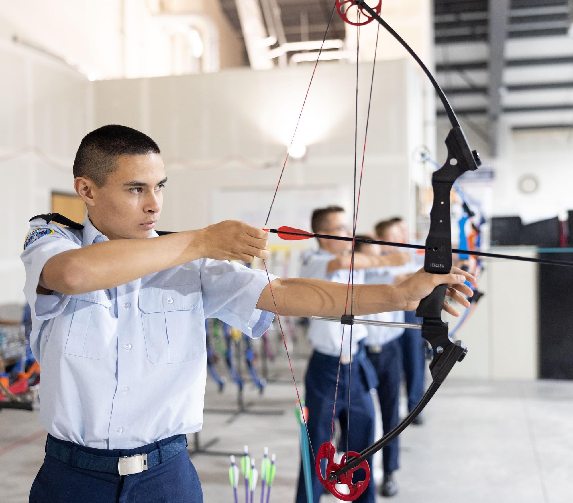 Cadet in AFJROTC uniform drawing compound bow during archery practice in indoor range