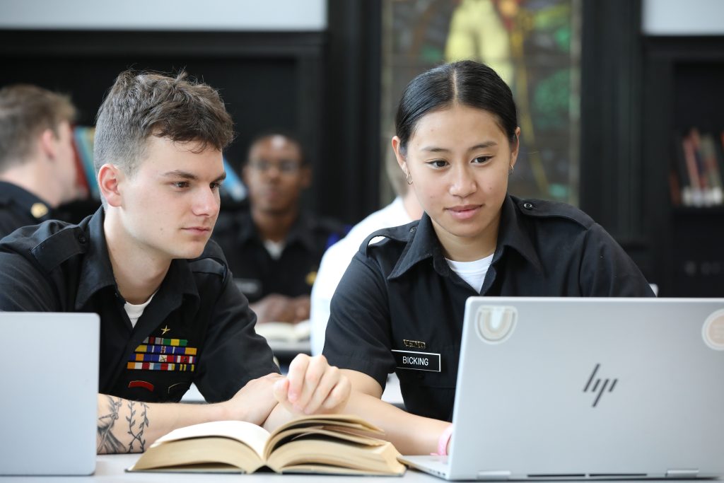 Two cadets in black uniforms studying together with laptops and textbook in library