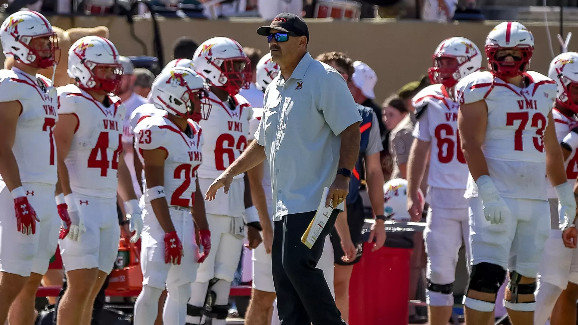 VMI football coach on the sideline with players in white uniforms with red VMI lettering during a game