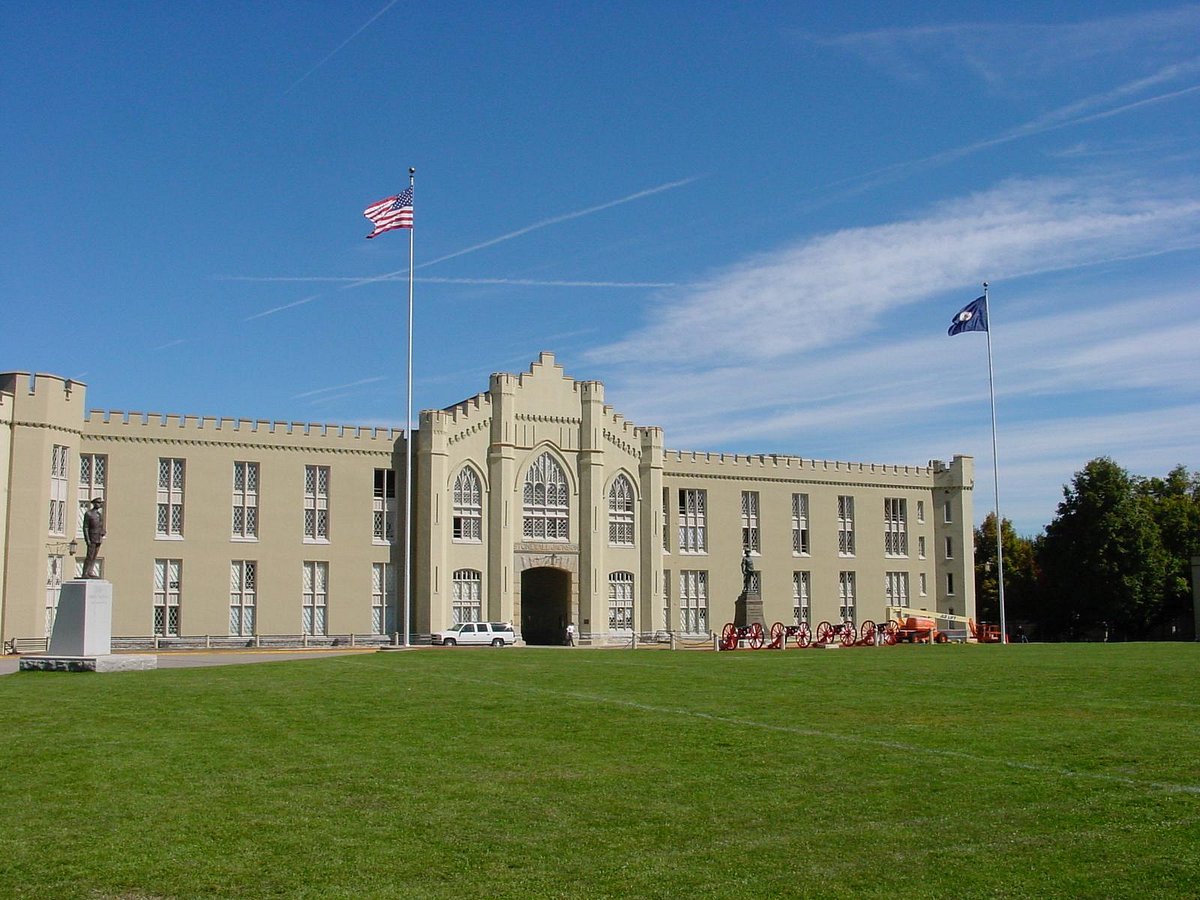 Front view of VMI's Gothic Revival limestone barracks with crenellated walls, American flag, Stonewall Jackson statue, and historic red cannons on the parade ground