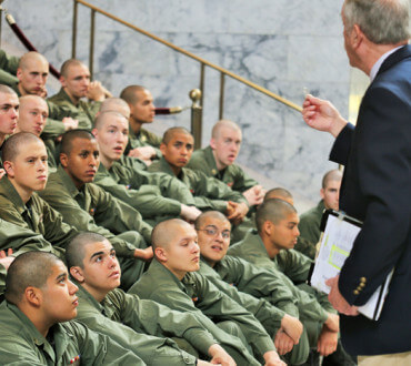 Cadets in olive uniforms seated on steps listening to speaker in suit at state capitol building