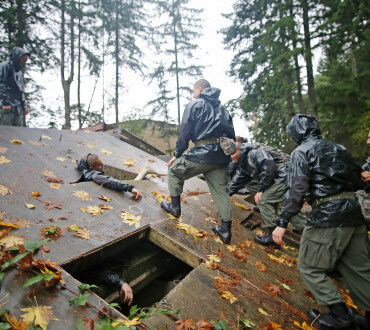 Cadets in camouflage and olive uniforms navigating wooden obstacle course structure in forest setting