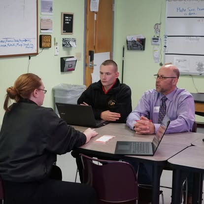 Cadet in black uniform meeting with instructor and staff at table with laptops in classroom