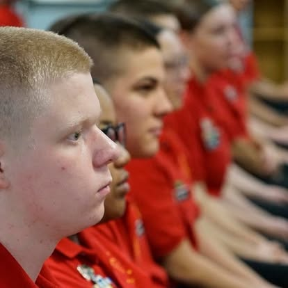 Row of cadets in red polo shirts with military-style haircuts seated attentively during assembly