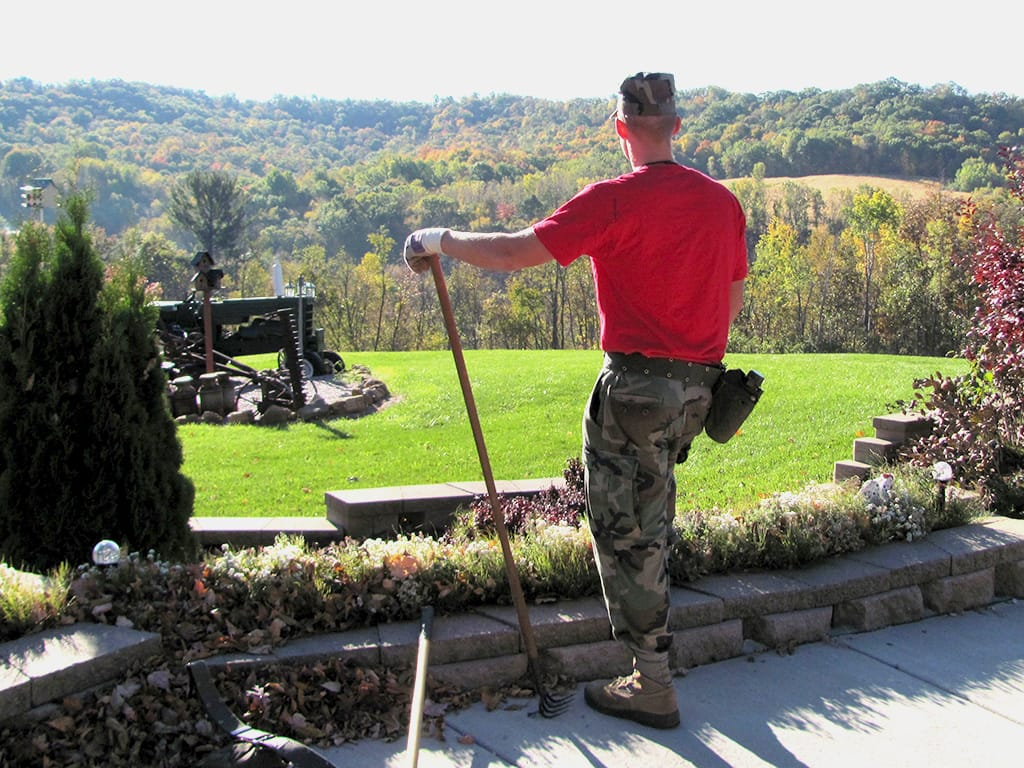 Cadet in red shirt and camo pants doing landscaping work overlooking autumn hillside at Fort McCoy